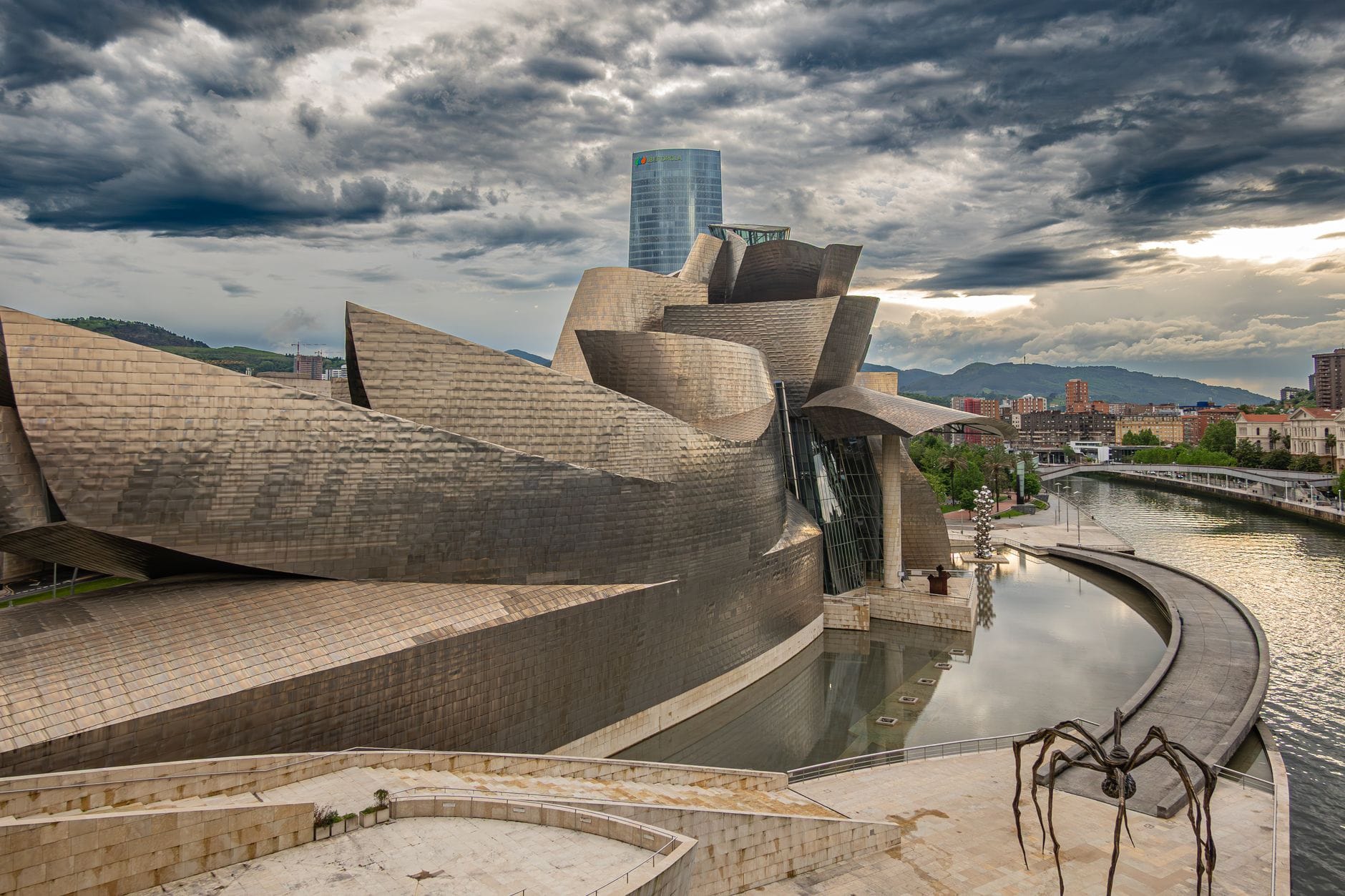 วิวกว้างของ Guggenheim Museum Bilbao พร้อมท้องฟ้าฤดูหนาวที่มีเมฆครึ้มอย่างน่าทึ่ง ริมแม่น้ำ และประติมากรรมรูปแมงมุมในเบื้องหน้า