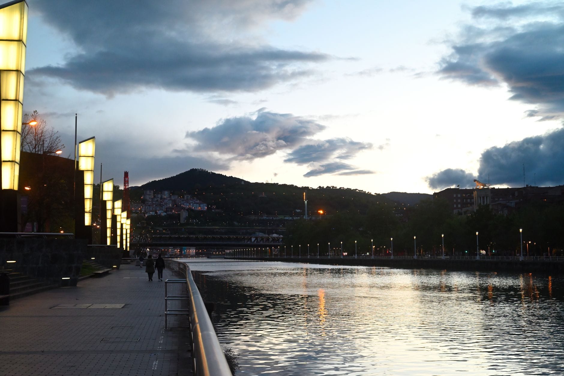 Vista nocturna del paseo fluvial de Bilbao con luces modernas, reflejos en el agua y personas paseando mientras el cielo transita del día a la noche.
