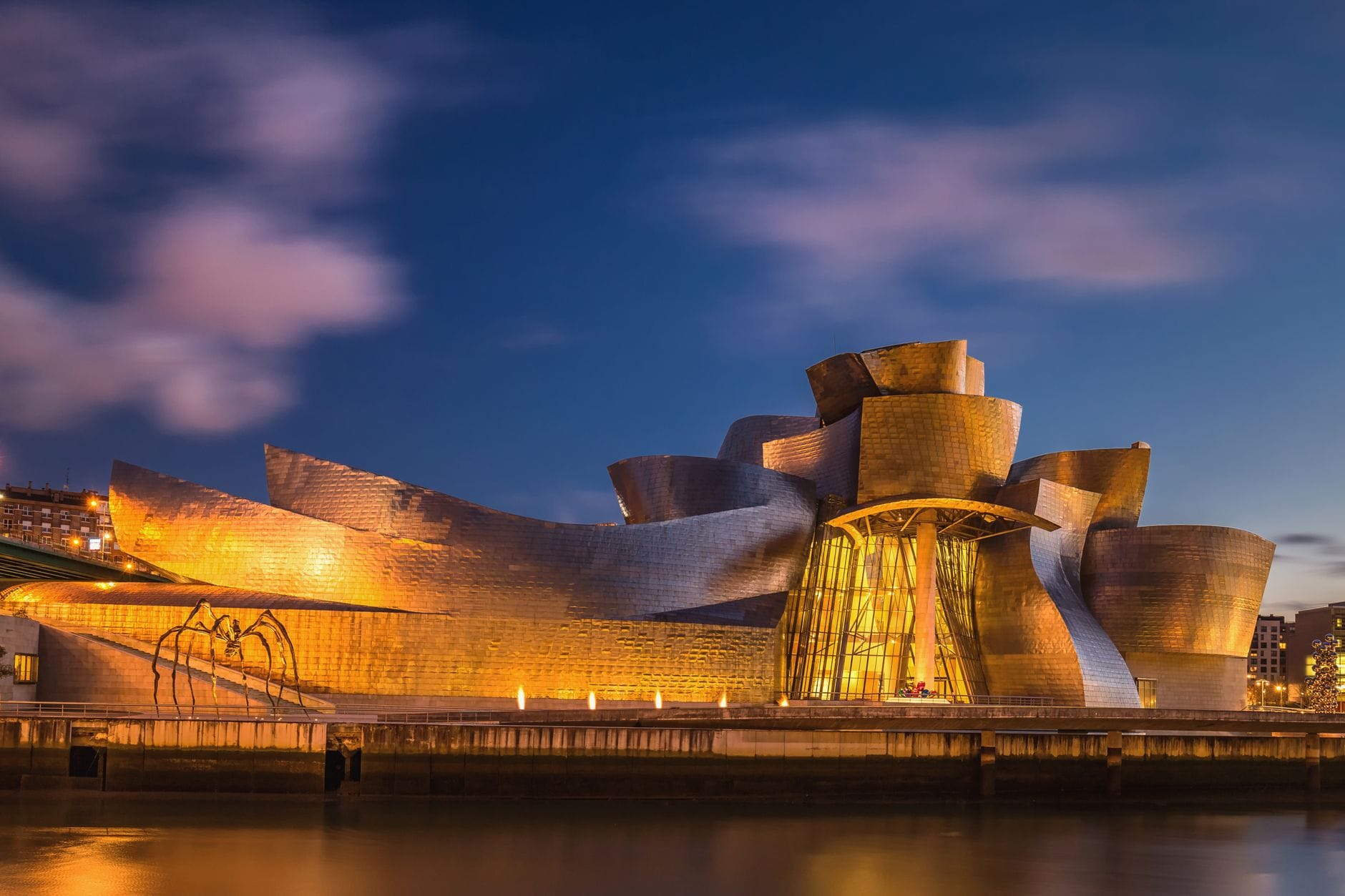 El Museo Guggenheim Bilbao iluminado bajo las luces de la noche, con reflejos en el río y un dramático cielo nocturno.