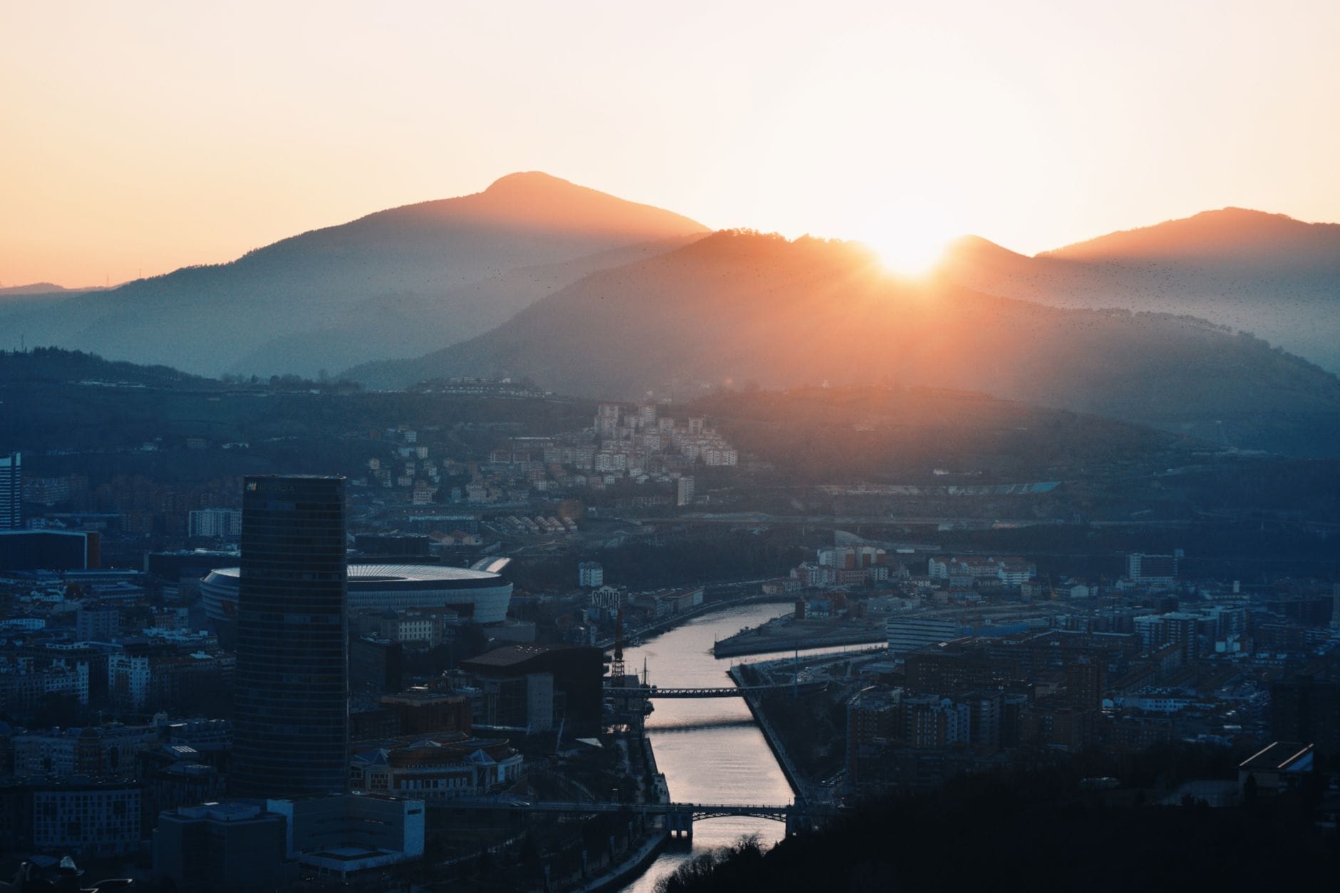 El sol poniente detrás de las colinas sobre Bilbao con paisaje urbano, río y cielo resplandeciente, creando una atmósfera de crepúsculo dorado sobre la ciudad.