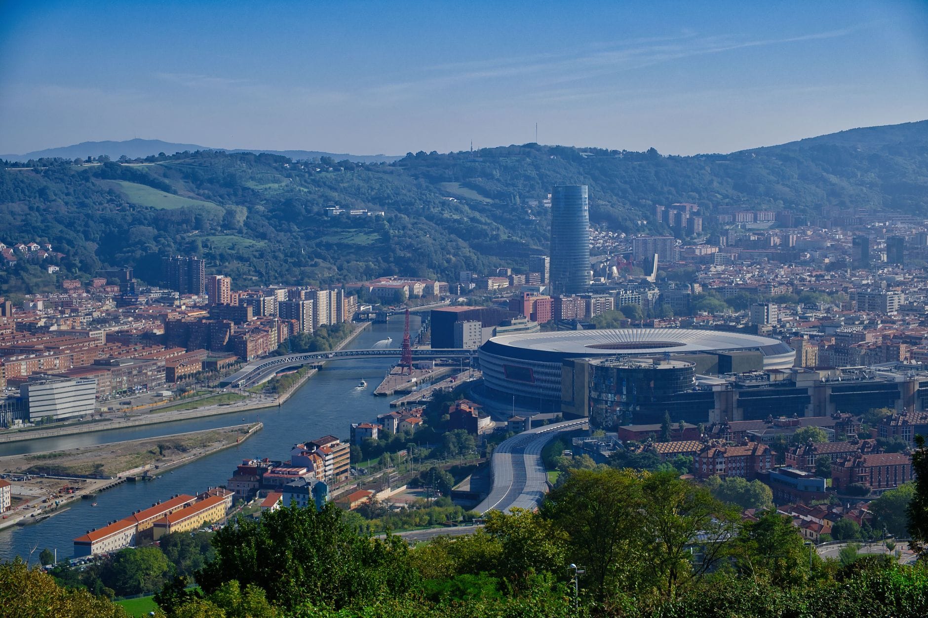 A panoramic cityscape showing the Bilbao river, iconic buildings, green hills, and a mix of modern and historic architecture under a blue sky.