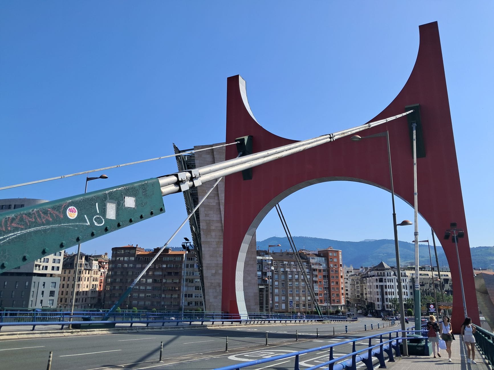 Wyraźny czerwony łuk Mostu La Salve w pobliżu Muzeum Guggenheim w słoneczny dzień, z panoramą Bilbao w tle