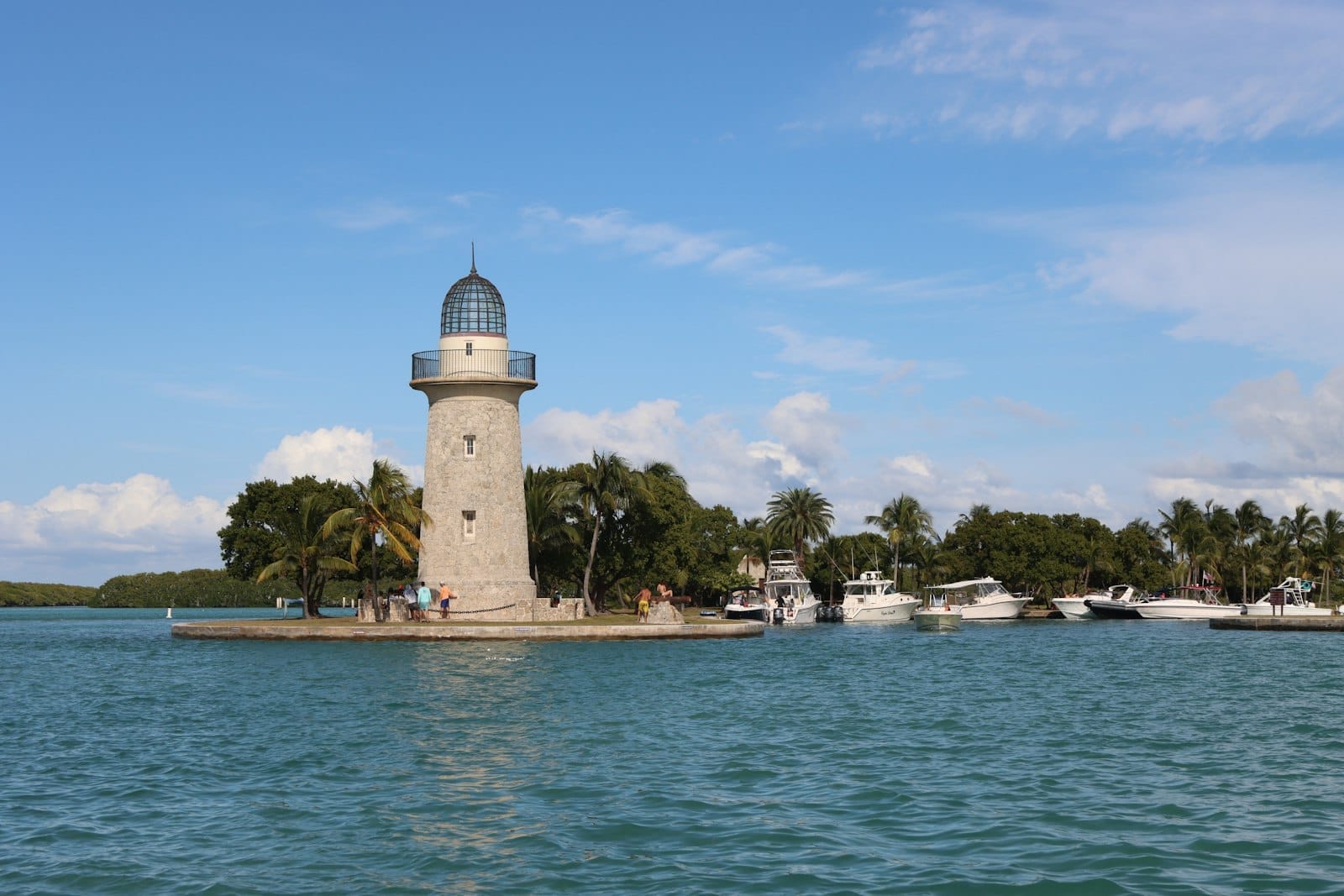 Faro in pietra sulla riva del Biscayne National Park, circondato da palme e barche su acque turchesi sotto un cielo azzurro e soleggiato.