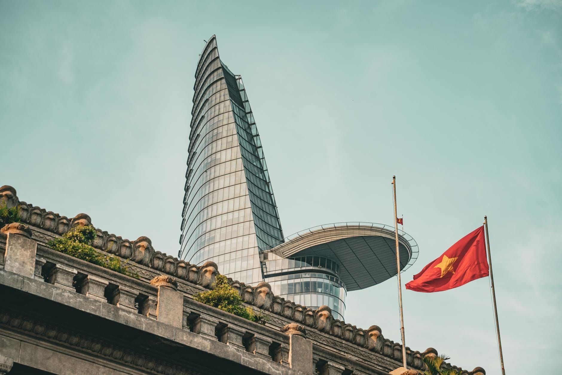 La Torre Financiera Bitexco se eleva sobre un edificio antiguo con una bandera vietnamita, enmarcada contra un cielo azul despejado en el centro de Ho Chi Minh City.
