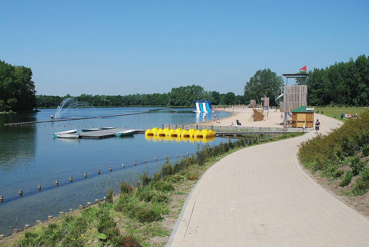 Sunny view of Blaarmeersen Sports and Recreation Park with a sandy beach, lake, pedal boats, water slides, and green trees under a clear blue sky.