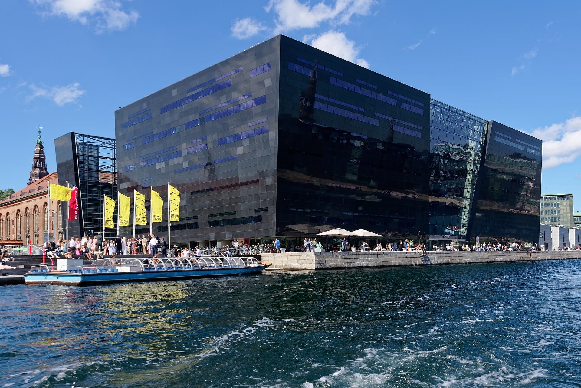 Il moderno edificio della biblioteca Black Diamond con facciata nera riflettente, persone sul lungomare, bandiere gialle e una barca sul canale sotto un cielo azzurro limpido a Copenaghen.