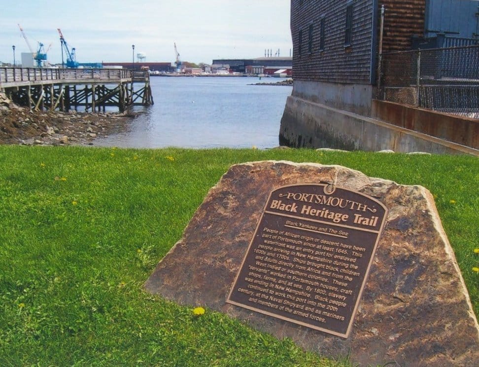 Una targa in bronzo del Portsmouth Black Heritage Trail montata su un masso, con prato, lungomare, molo in legno ed edifici industriali sotto un cielo azzurro.