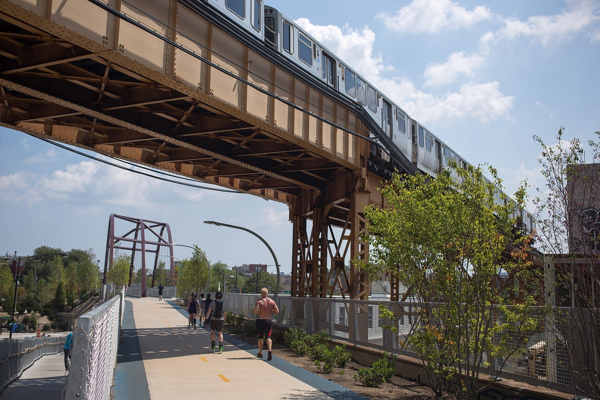 Persone che fanno jogging e camminano sul Bloomingdale Trail (The 606) sotto un treno sopraelevato di Chicago che passa in un pomeriggio soleggiato.