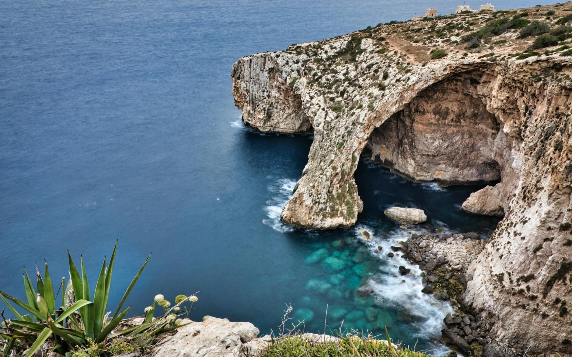 Vista panoramica della Grotta Azzurra di Malta con le imponenti scogliere calcaree, un grande arco marino, l'acqua turchese e la vegetazione costiera in primo piano.