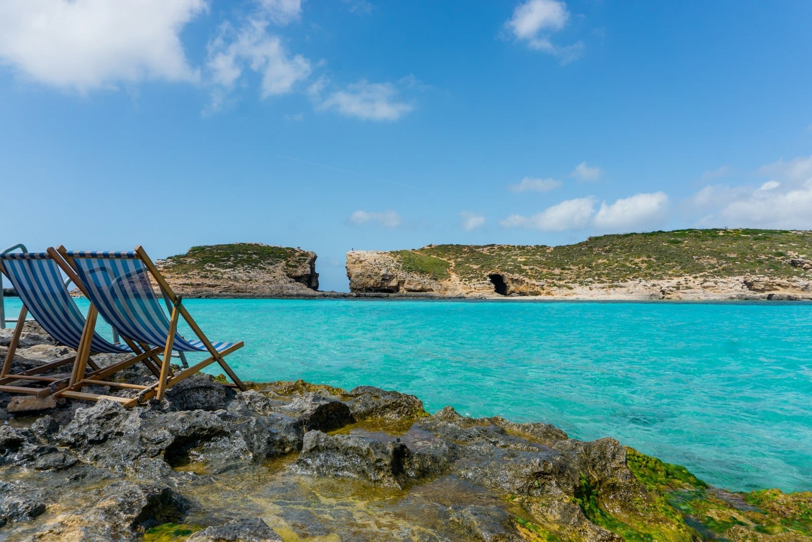 Due sedie a sdraio a righe blu su una riva rocciosa con vista sulle acque turchesi e le scogliere frastagliate della Blue Lagoon, Comino, sotto un cielo luminoso.