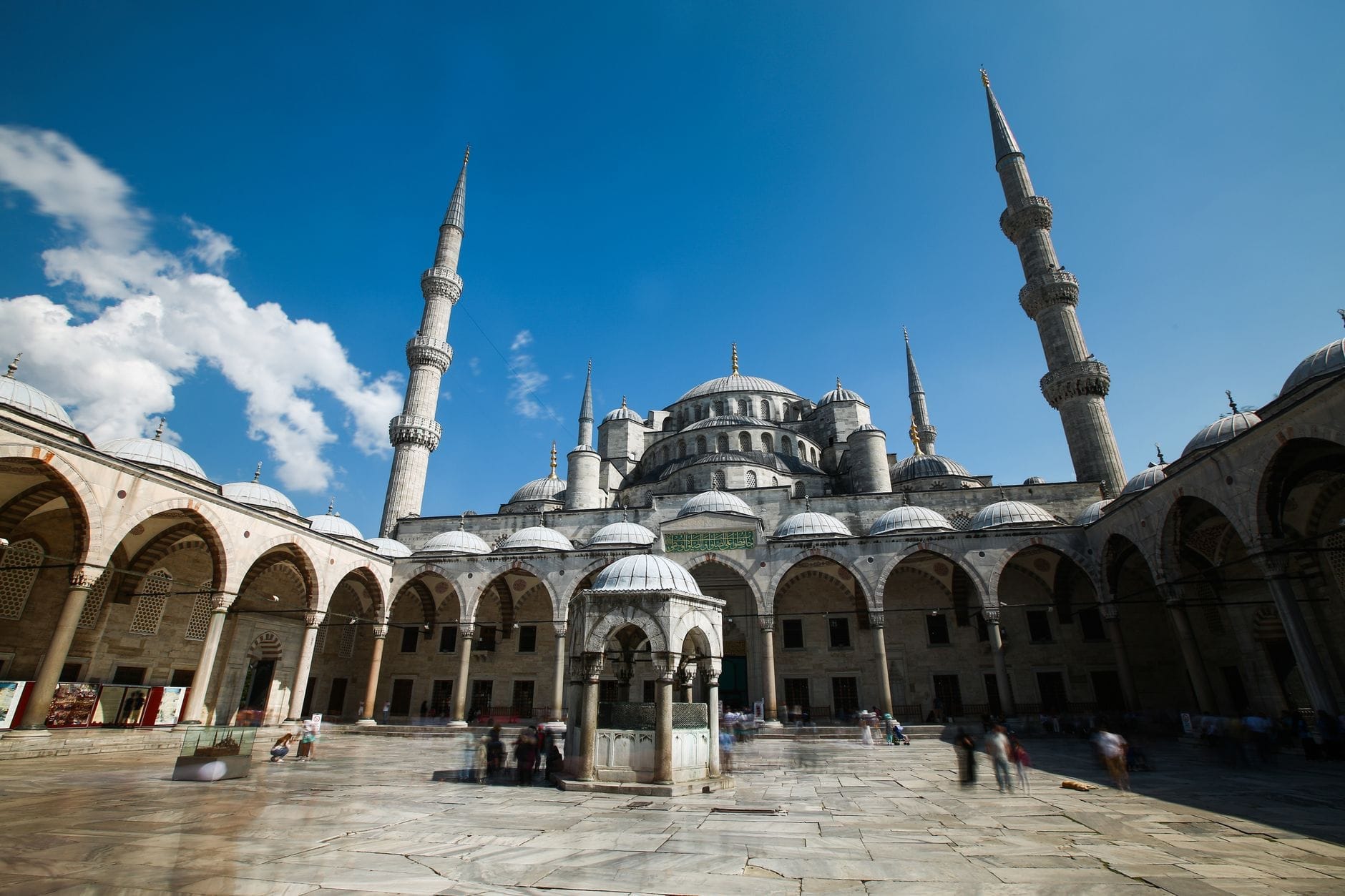 Vista grandangolare del cortile della Moschea Blu e delle cupole principali sotto un cielo azzurro intenso, che mettono in risalto la grandiosità dell'edificio e il suo ingresso accogliente.