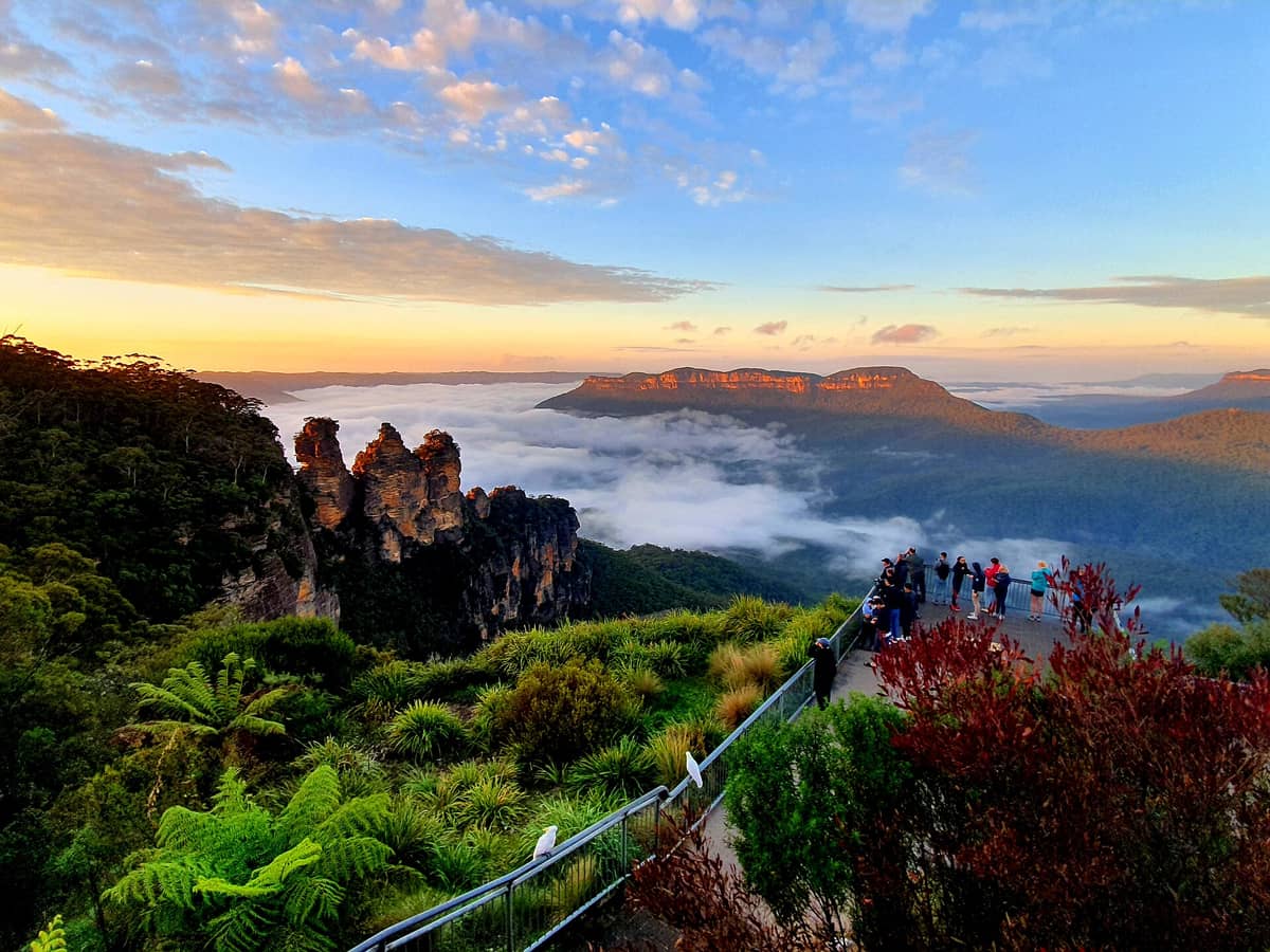 Aussichtspunkt Echo Point mit Touristen, die bei Sonnenaufgang die Felsformation der Three Sisters und das Jamison Valley in den Blue Mountains betrachten, mit üppigem Grün und Nebel im Tal.