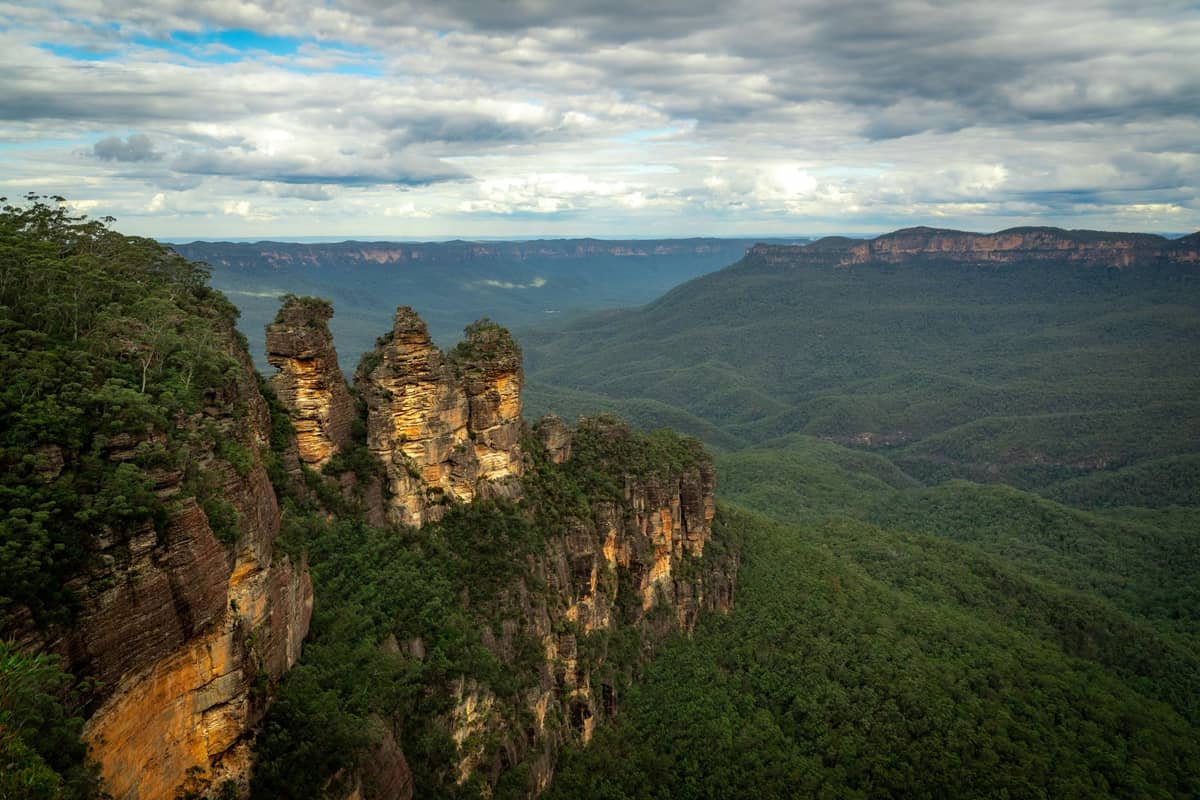 Landschaftsblick auf die Felsformation Three Sisters und das üppige Tal der Blue Mountains unter einem teilweise bewölkten Himmel.