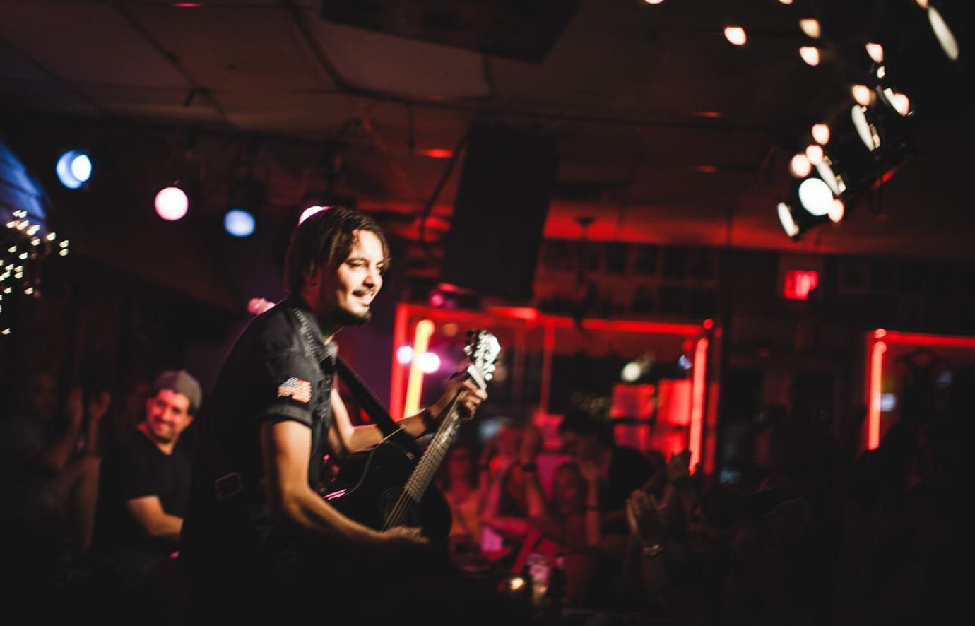Músico actuando con una guitarra acústica en el Bluebird Cafe en Nashville, rodeado de cálida iluminación escénica y un público íntimo.