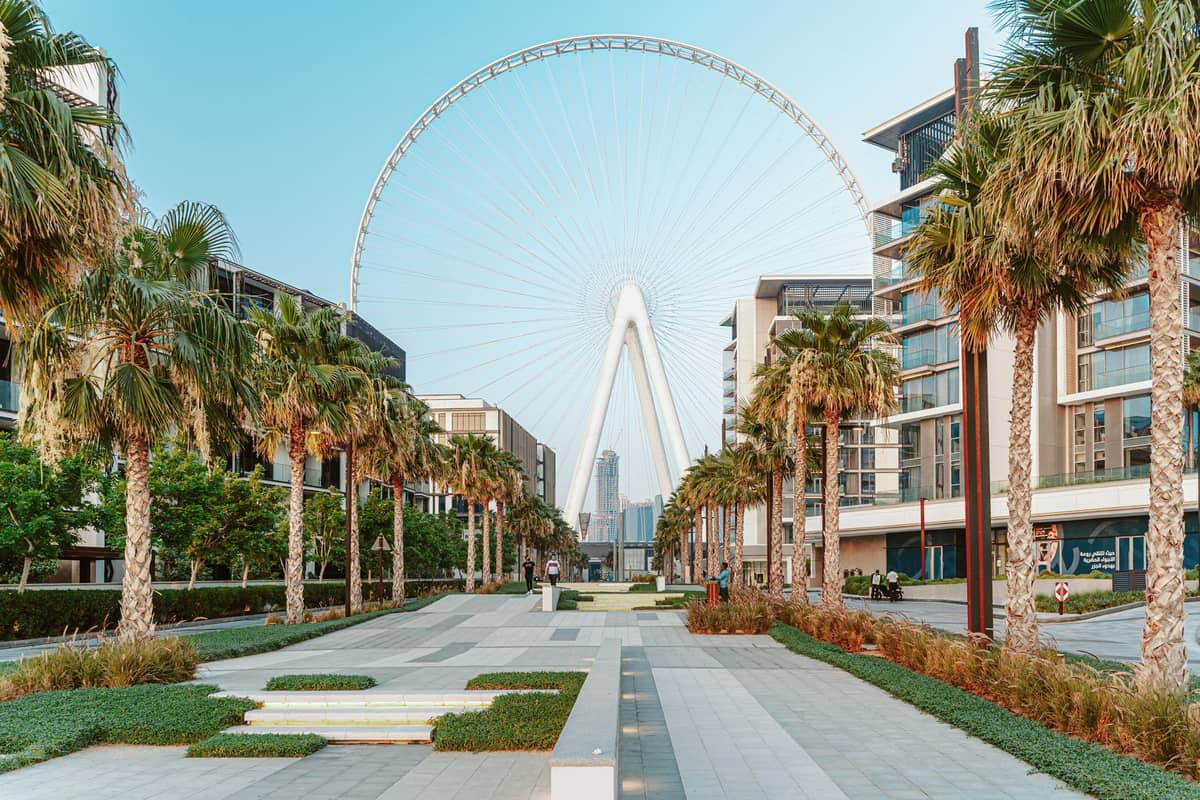 Breite, mit Palmen gesäumte Fußgängerpromenade auf Bluewaters Island in Dubai, die zu Ain Dubai, dem höchsten Riesenrad der Welt, führt, mit modernen Gebäuden auf beiden Seiten.