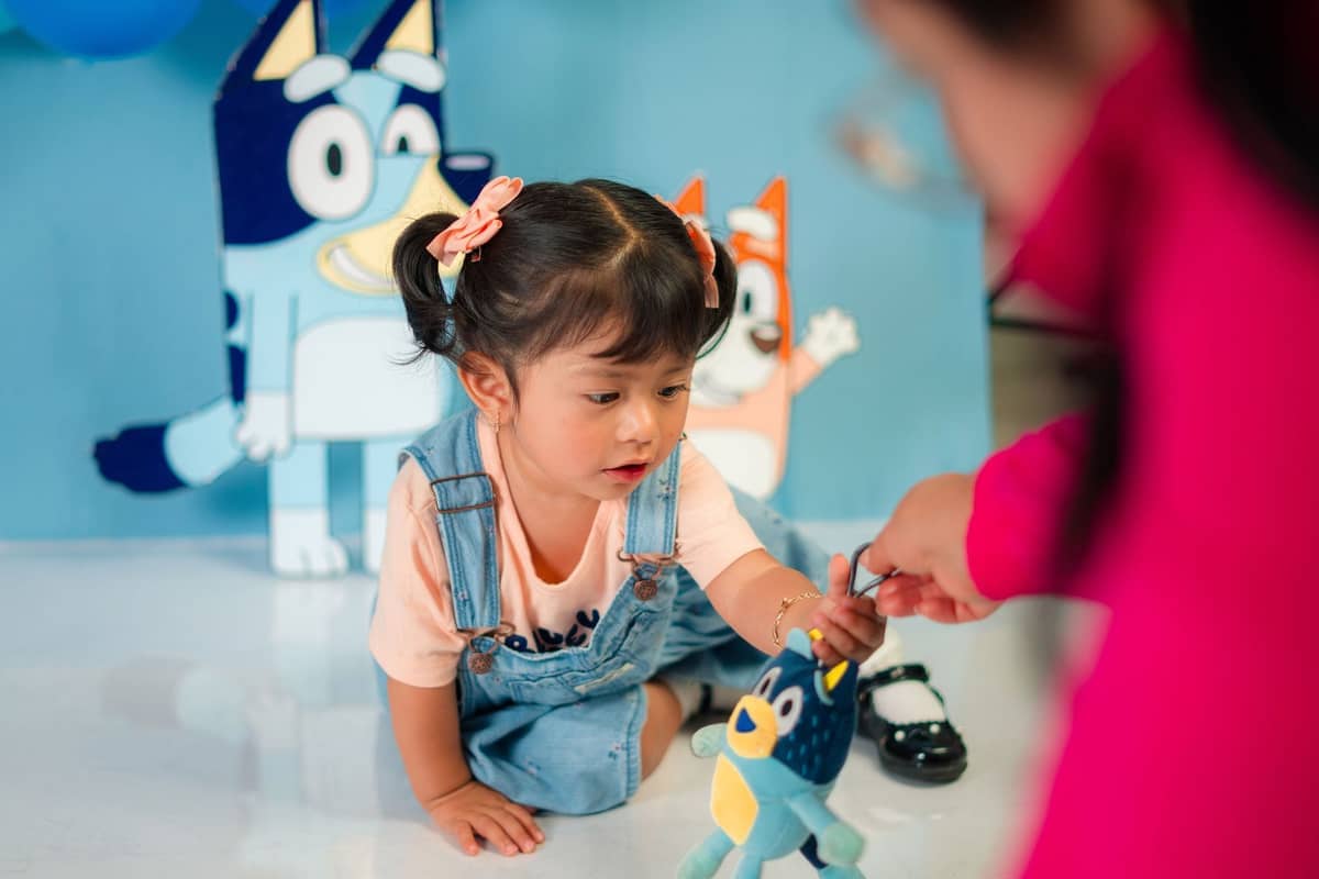 Un niño pequeño sentado en el suelo en Bluey's World en Brisbane, alcanzando un peluche de Bluey con recortes de dibujos animados detrás.