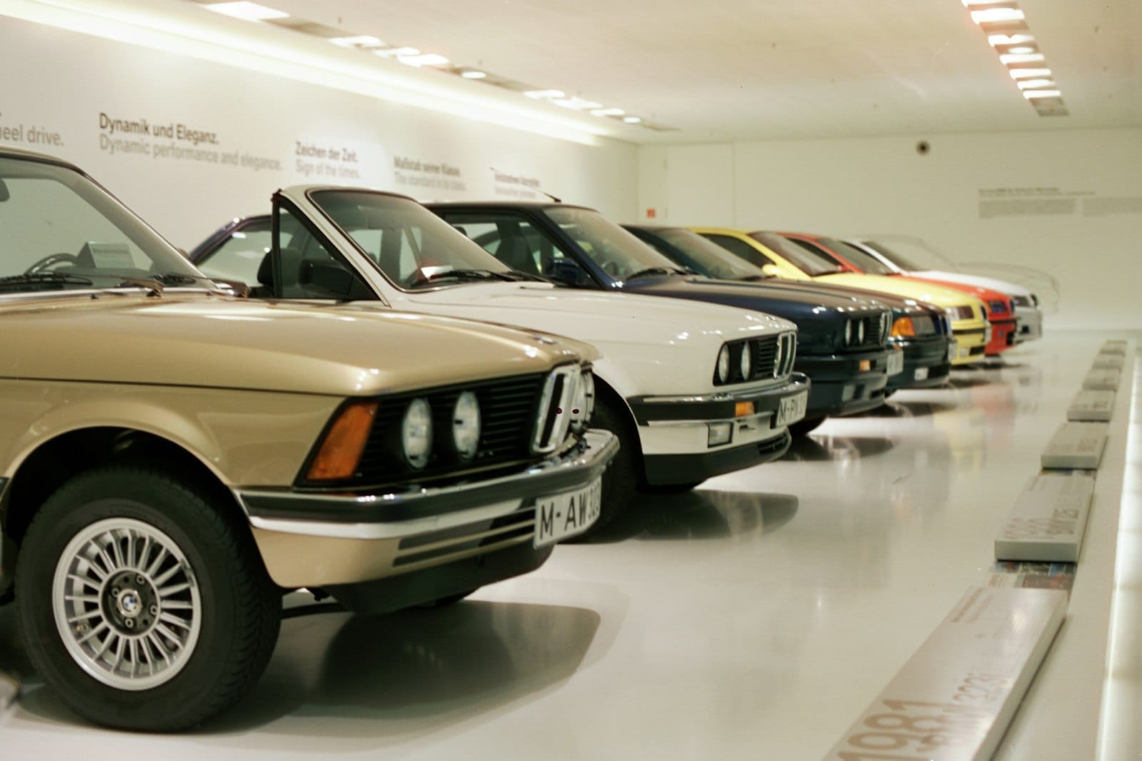 แถวรถยนต์ BMW คลาสสิกที่จัดแสดงภายใต้แสงไฟสว่างใน BMW Museum München โดยมีข้อมูลไทม์ไลน์ที่มองเห็นได้บนพื้น