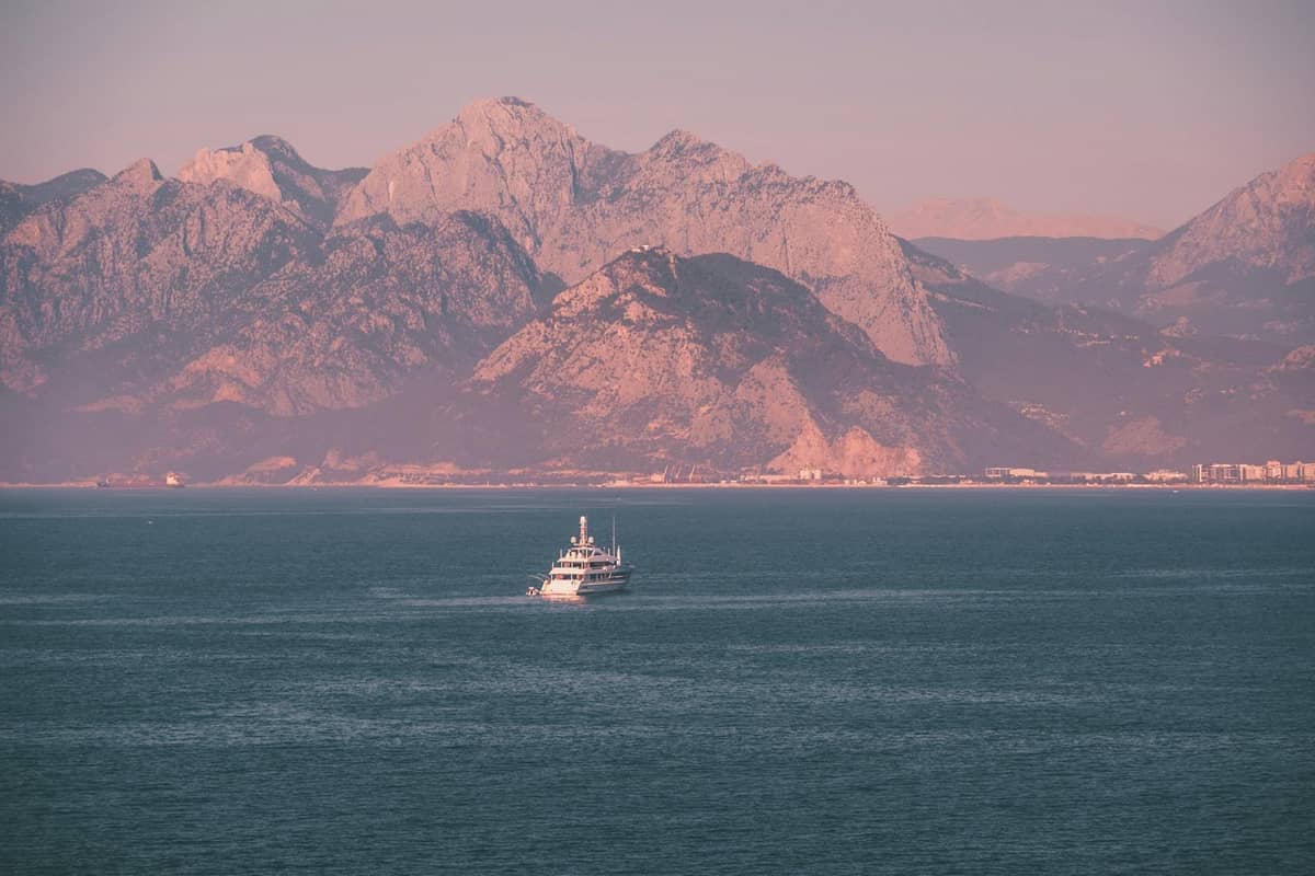 A boat cruising on the Mediterranean Sea with Antalya's dramatic mountains and city skyline in the background at sunset.