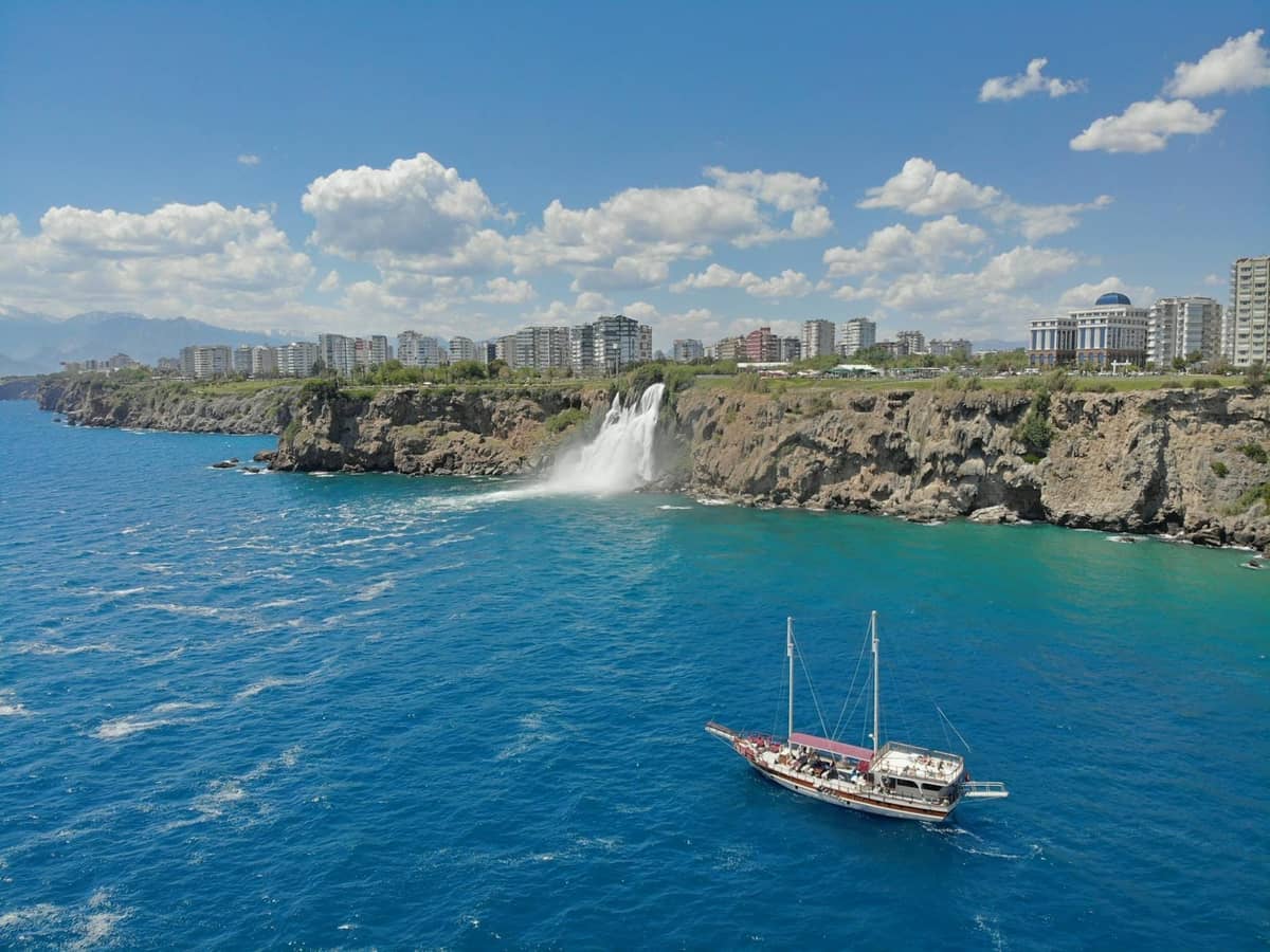 Boat sailing near Antalya’s dramatic coastal cliffs with waterfalls and cityscape visible in the background under a blue sky.