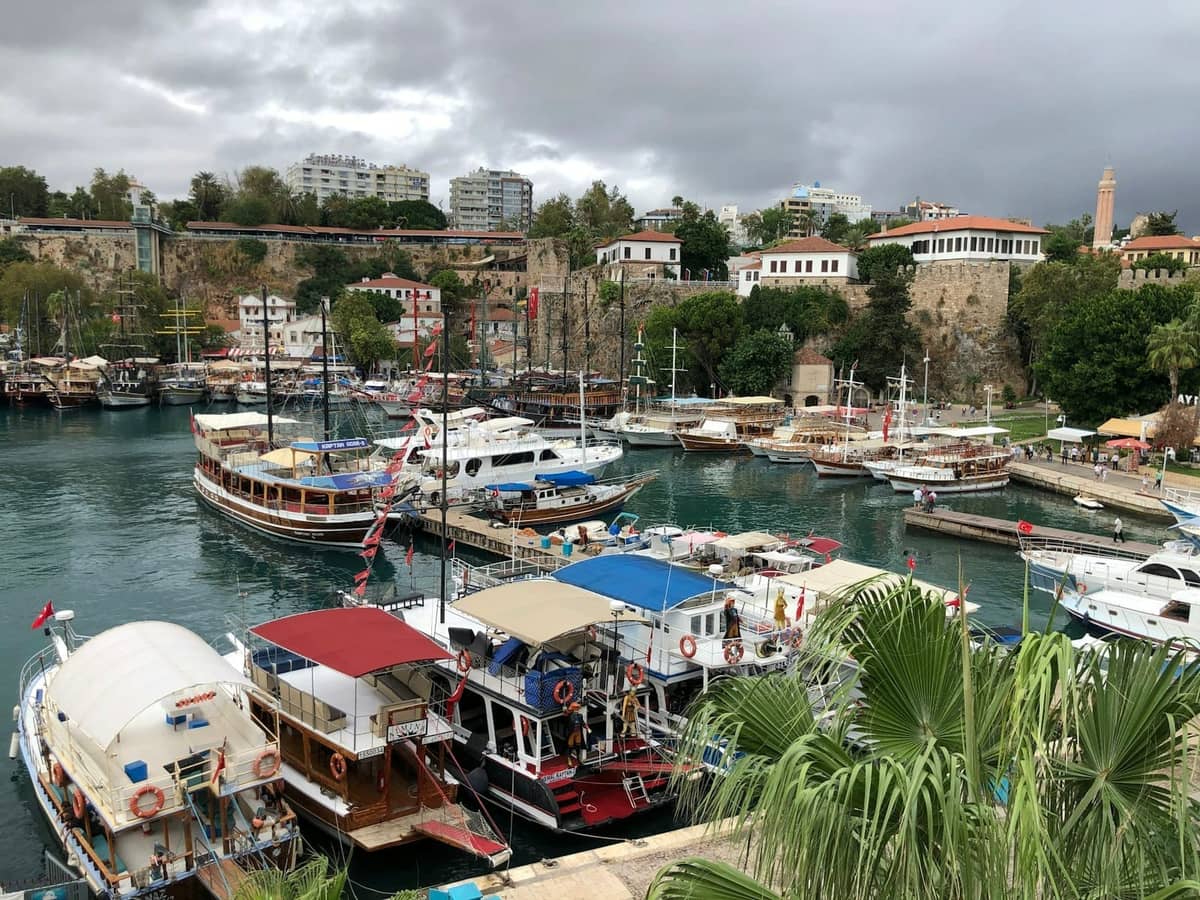 Central Antalya harbor with tour boats docked, historic stone walls, lush greenery, and city buildings in the background.