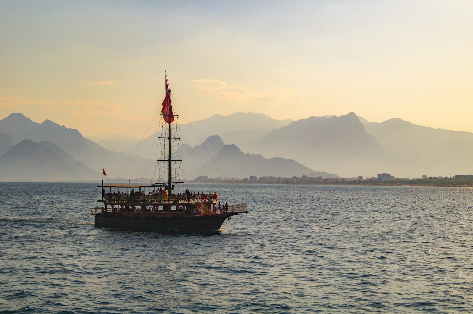 Barco tradicional navegando pelo mar azul ao pôr do sol com as montanhas de Antalya ao fundo sob um céu quente.