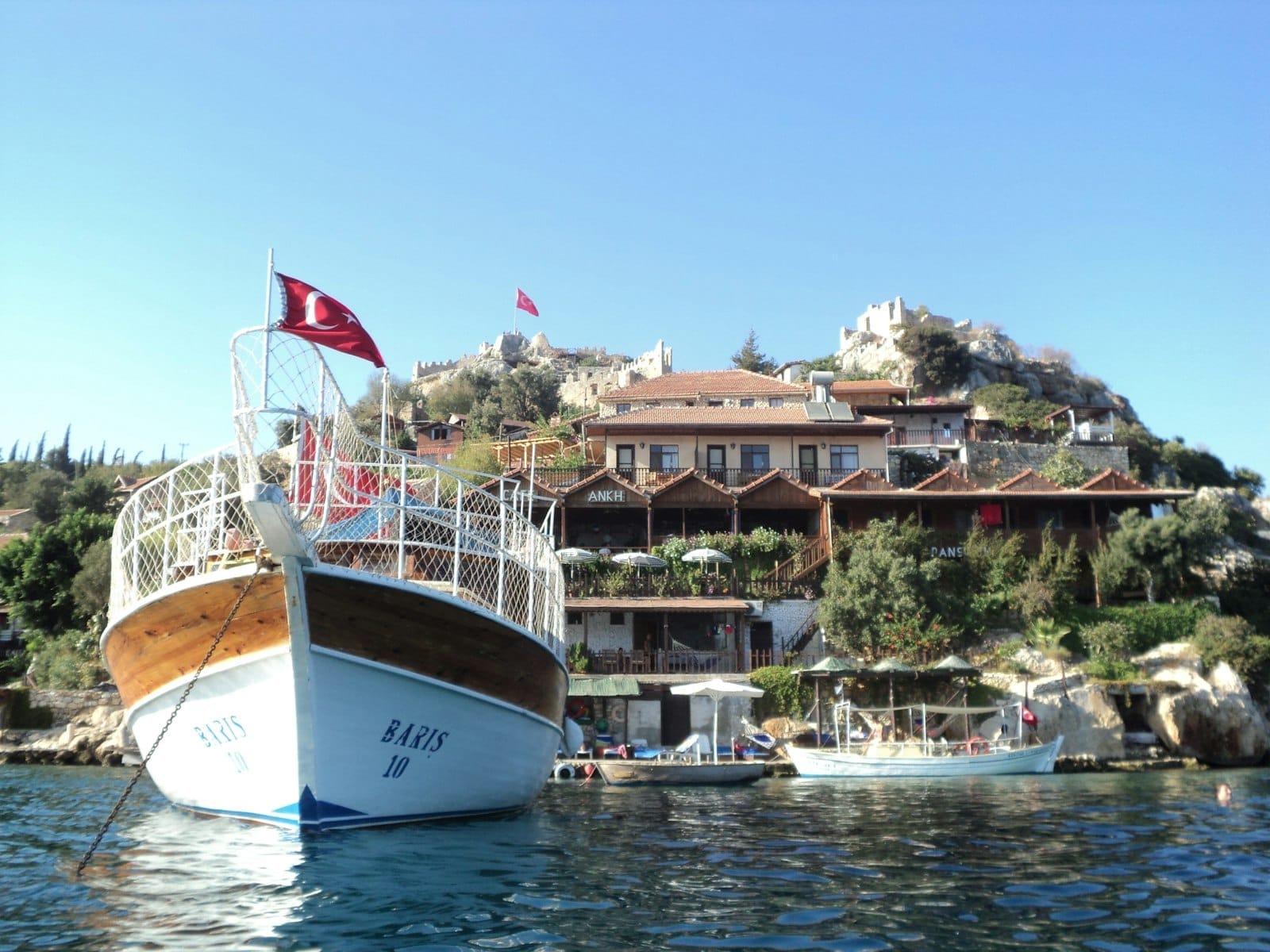 Una barca turistica in legno con bandiera turca ancorata presso il Kaleiçi Marina, con ville sul versante collinare e rovine antiche sotto un cielo blu limpido.