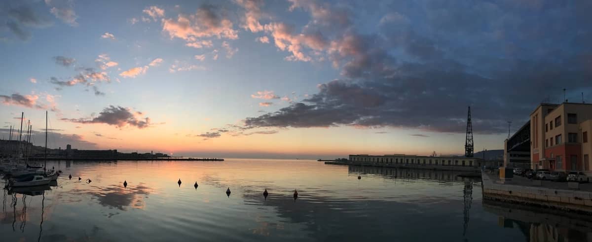 Weiter Blick auf die Uferpromenade bei Sonnenaufgang mit ruhigem Meer, kleinen Booten, Altstadtgebäuden auf der rechten Seite und dem Matejuška-Steg in Split.