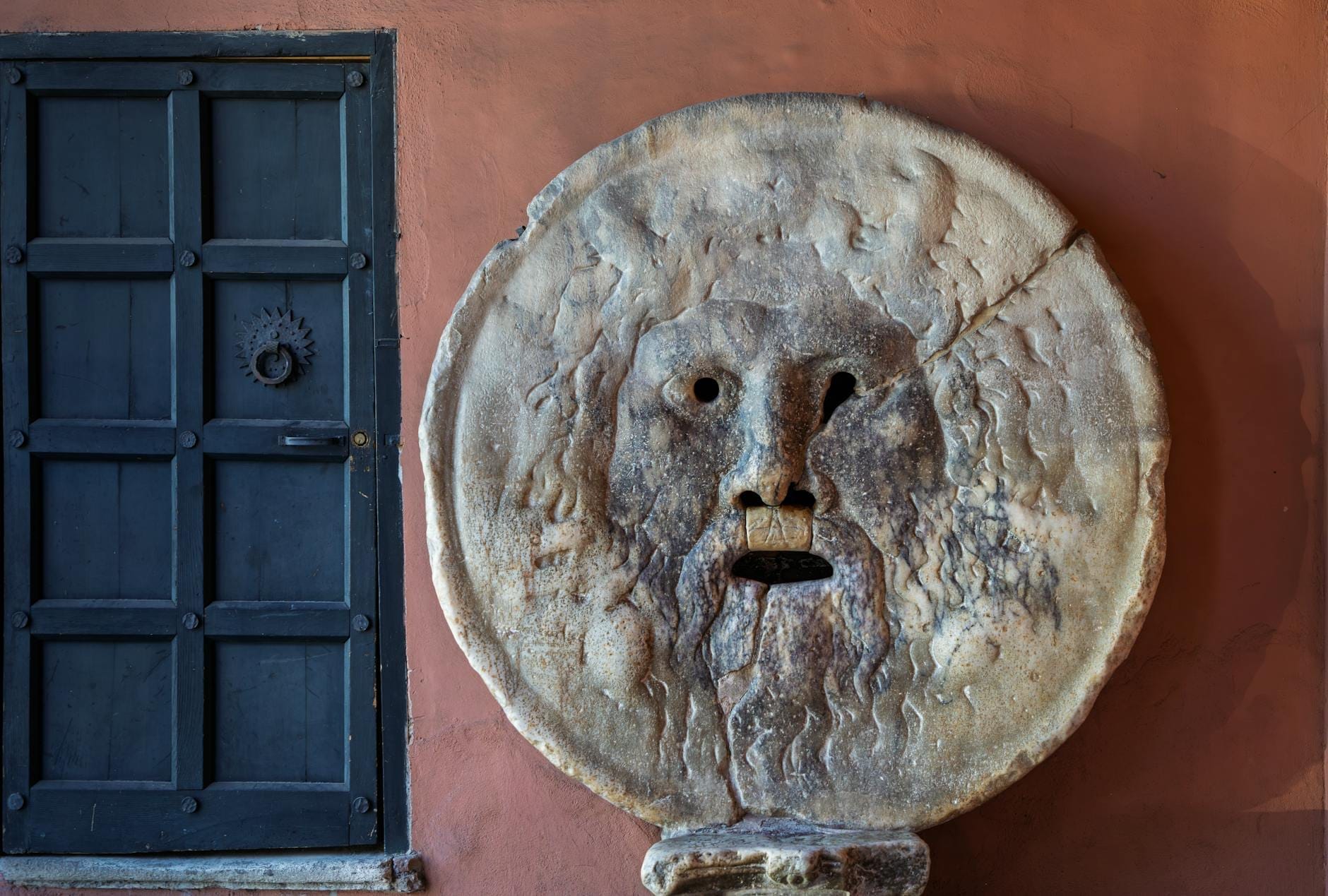 Close-up view of the Bocca della Verità, a large ancient marble mask with a carved face, set into a wall next to a dark door.