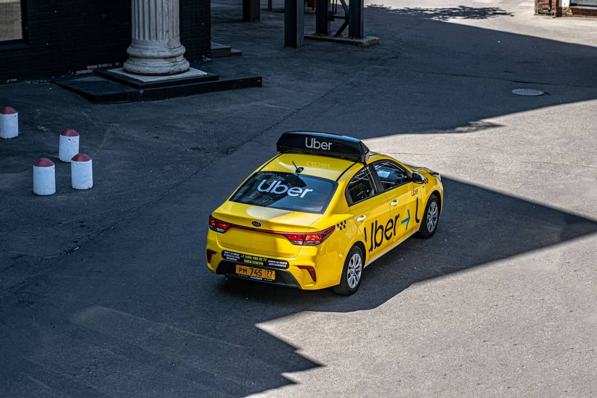 A yellow car with prominent Uber branding driving on an urban street in daylight.