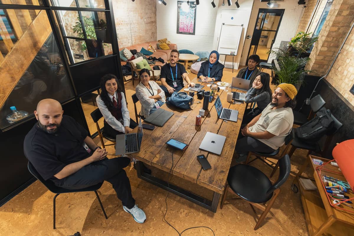 Group of people working on laptops around a large wooden table in a modern coworking space with exposed brick walls.