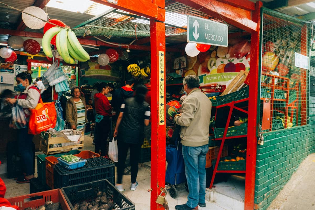 A lively local market in Bogotá with people shopping for fruits and produce at various colorful stalls under a red awning.