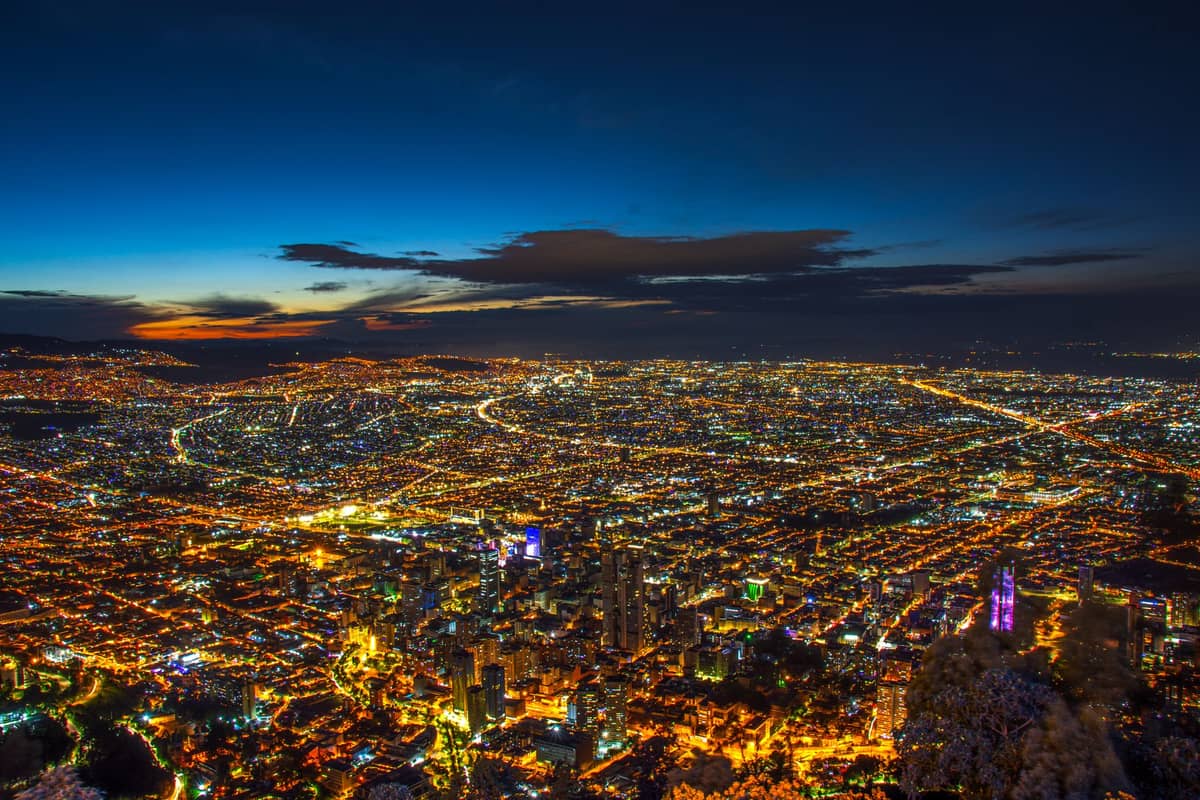 A sweeping aerial view of Bogotá at night, with the city lights stretching to the horizon and the Andean mountains in the background.