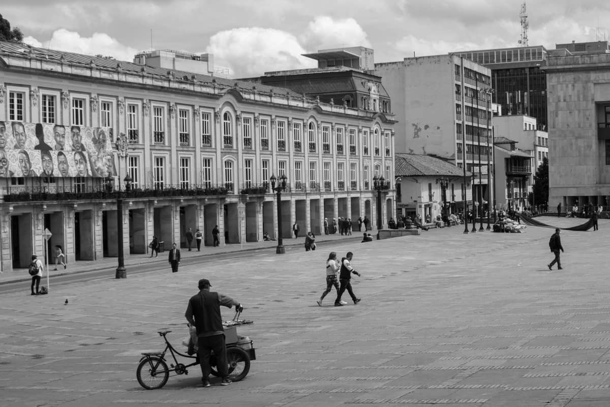Black and white photo of Bogotá's Plaza de Bolívar with neoclassical government buildings, pedestrians walking, and a person with a bike in the foreground.