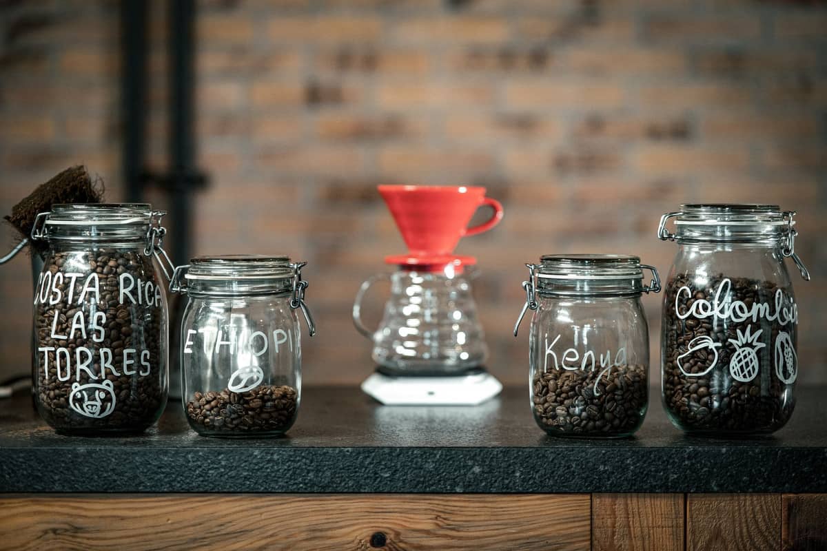 Glass jars with coffee beans labeled by country, including Colombia, on a cafe counter with a pour-over dripper in the background.