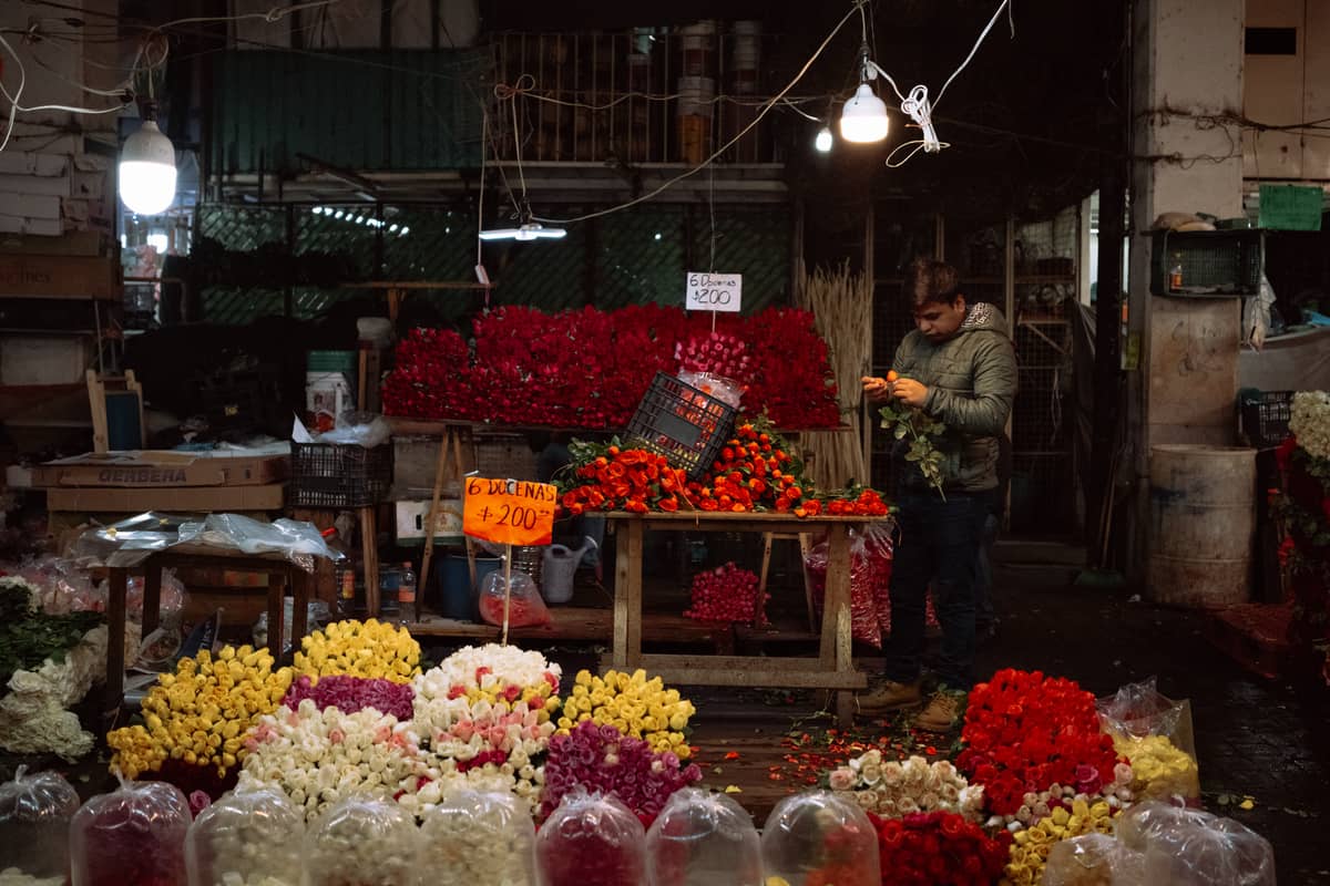Indoor flower stall with a man arranging bright bouquets among fresh roses and carnations at a bustling traditional market.