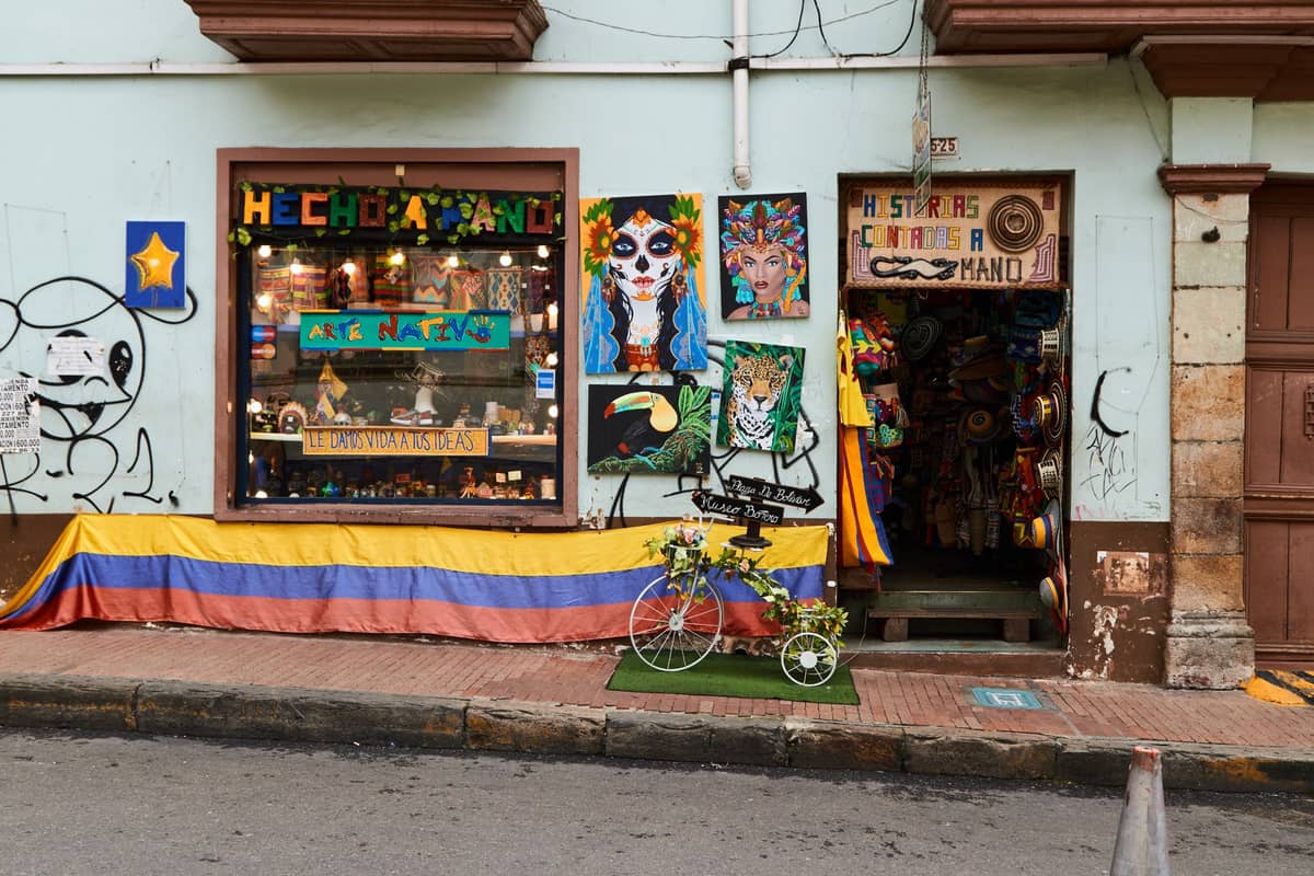 Street view of a colorful artisan craft shop with a Colombian flag, handmade items, and vibrant art lining the storefront in Bogotá.