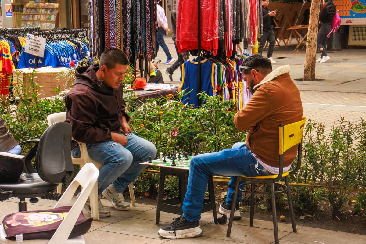 Two men playing chess at an outdoor market lined with colorful clothes and craft stalls.
