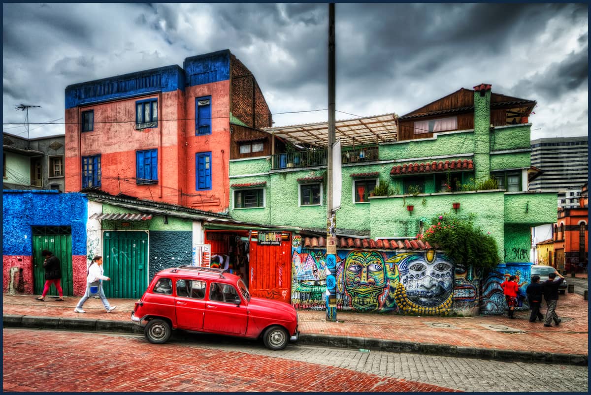 Colorful buildings with intricate street art murals in Bogotá’s La Candelaria neighborhood, with people walking on a brick-paved street and a red vintage car parked in front.