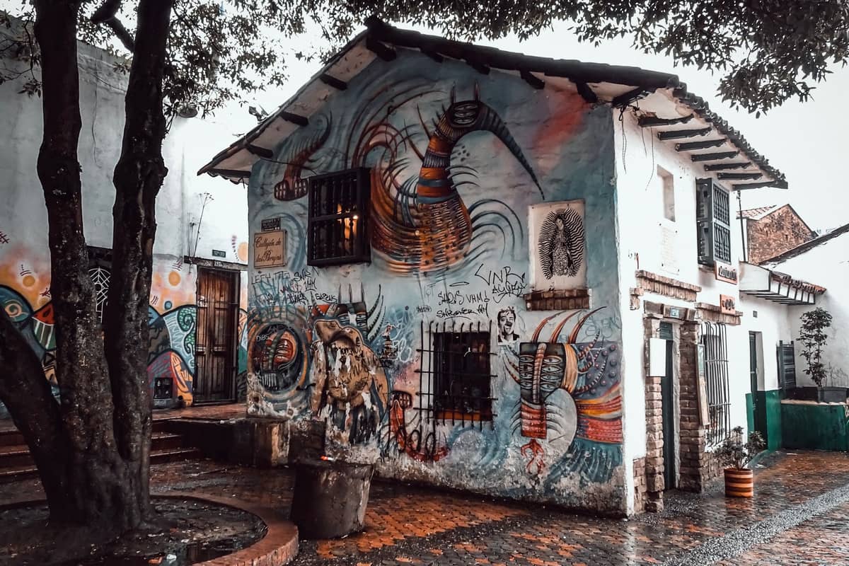 Colorful street art mural on a white building in Bogotá's La Candelaria, with wet cobblestone pavement and trees nearby.