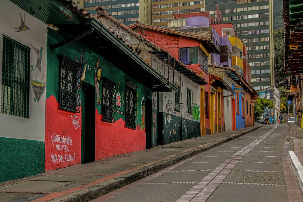 Colorful street in Bogotá's La Candelaria neighborhood with murals and painted houses, sloping uphill with city buildings in the background.