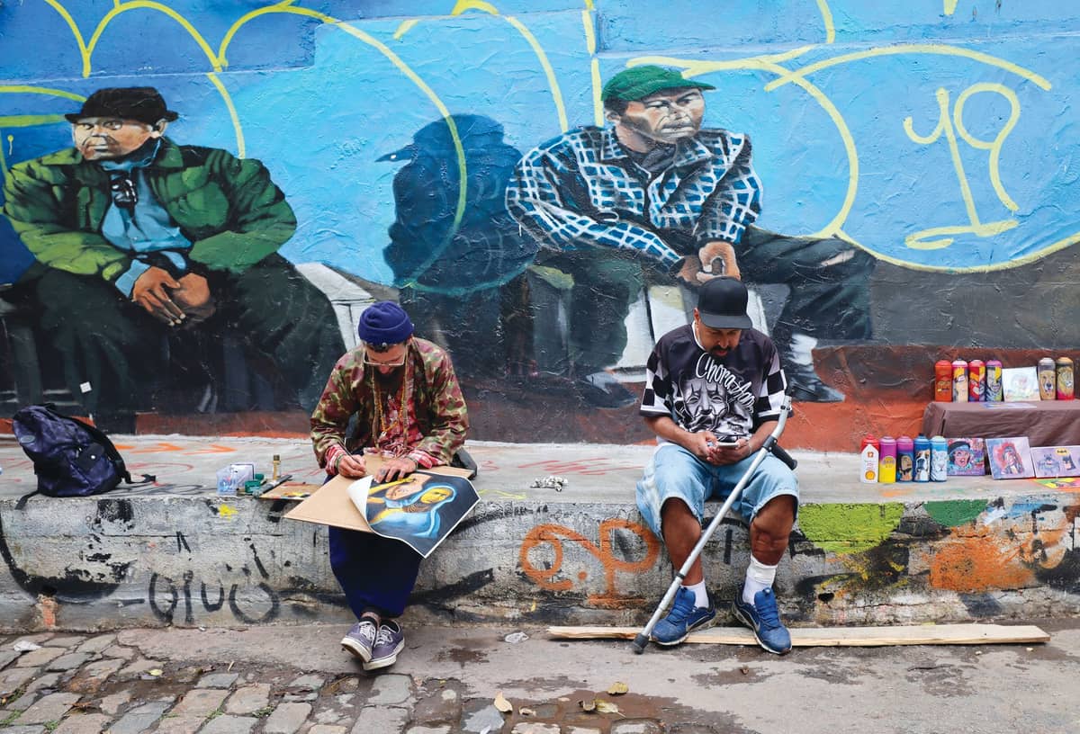 Two artists sit and work in front of a large colorful mural painted on a wall in Bogotá, surrounded by spray paint cans and art supplies.