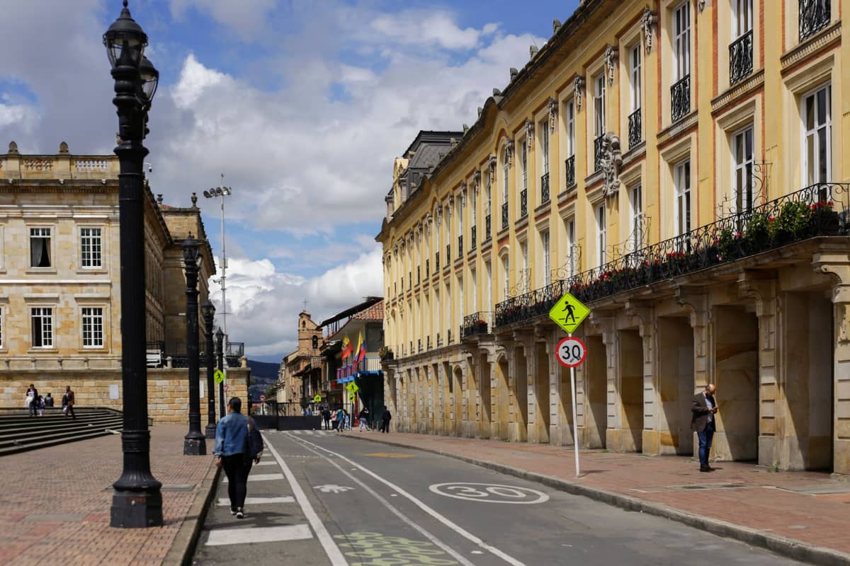 People walking along a wide street in Bogotá, Colombia, wearing layers and jackets with classic colonial buildings and cloudy sky.