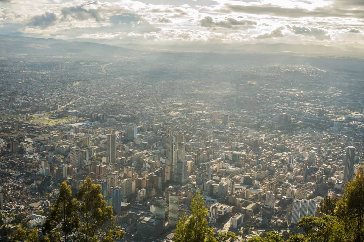 A panoramic, sunlit view over the city of Bogotá, Colombia, showing a mix of downtown skyscrapers, dense neighborhoods, and the surrounding Andean mountains.