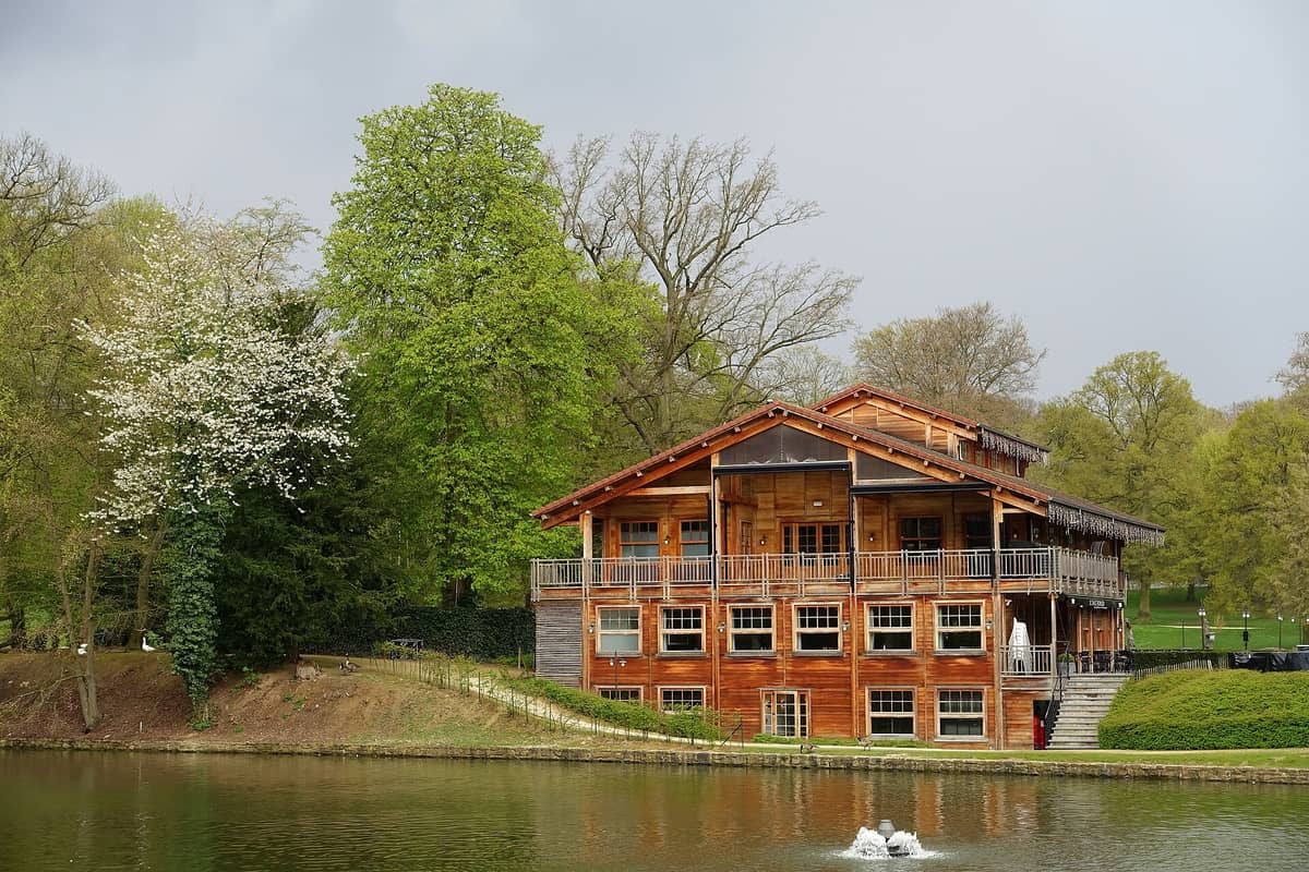 Grande restaurante de madeira estilo chalé à beira do lago central do Bois de la Cambre, cercado por árvores verdes e refletido na água.