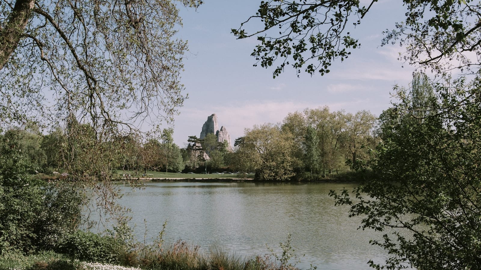 Lago tranquillo circondato da rigogliosa vegetazione e alberi nel Bois de Vincennes, con una struttura che ricorda un castello sullo sfondo sotto un cielo luminoso.