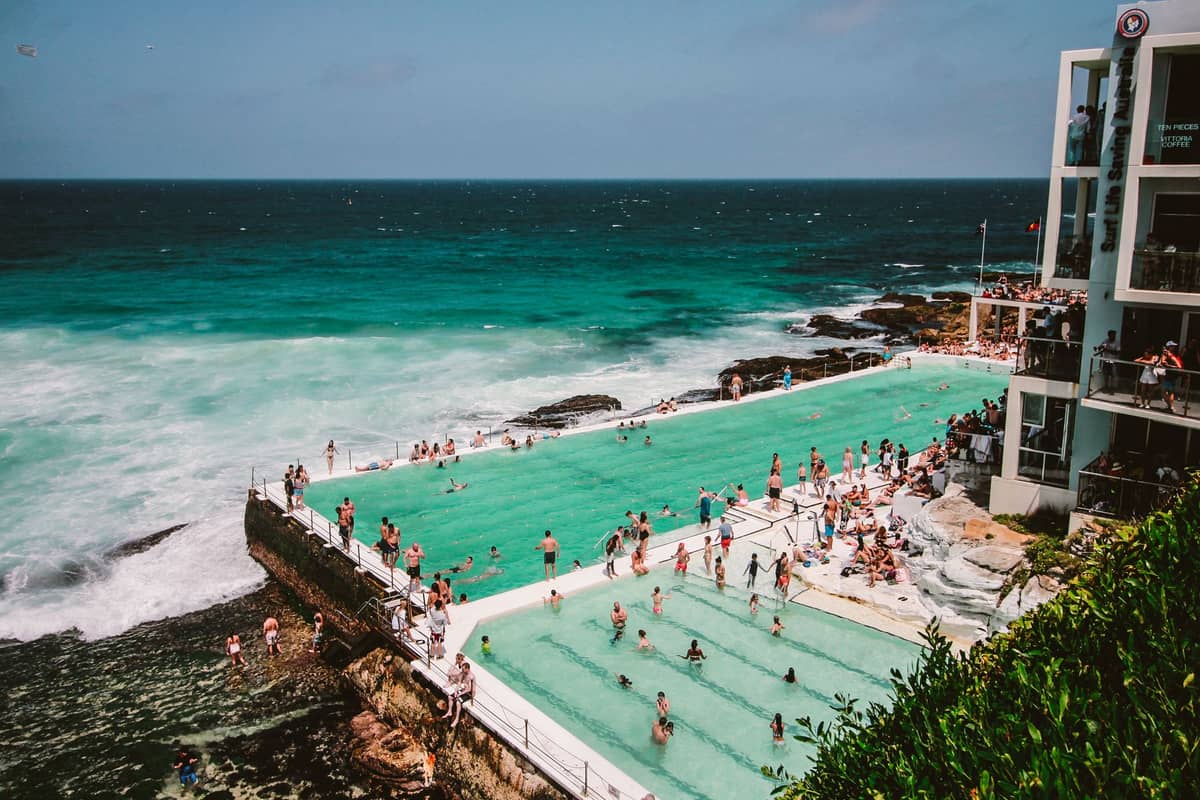 Panoramiczny widok na Bondi Icebergs Pool z pływakami w obu basenach, turkusowymi falami rozbijającymi się o krawędź i budynkiem klubu widocznym po prawej stronie.