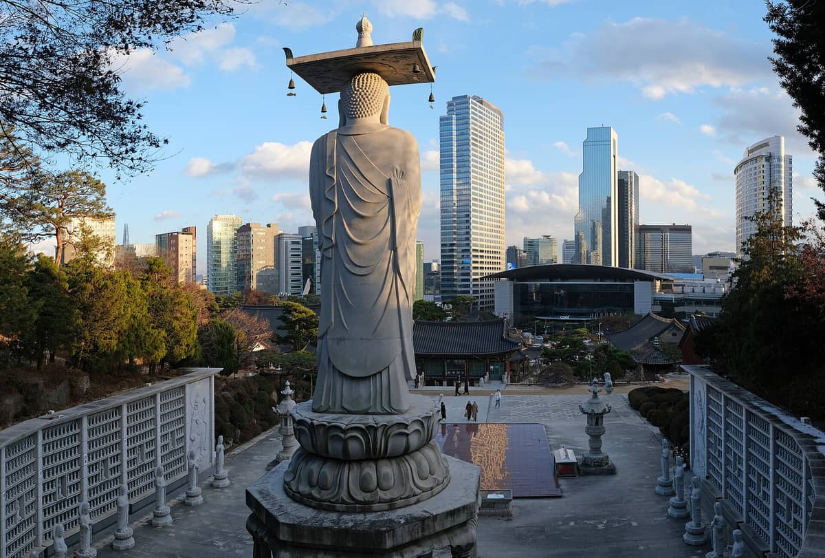 Vista traseira da grande estátua de Buda no Templo Bongeunsa com os edifícios tradicionais do templo e os arranha-céus modernos de Gangnam, Seul, ao fundo em um dia claro.