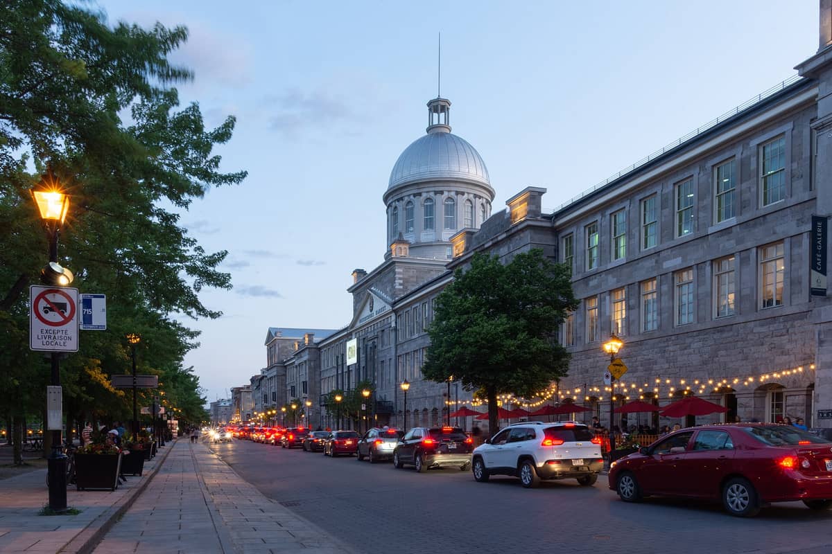 Der Bonsecours Market in Alt-Montréal in der Abenddämmerung, mit seiner markanten silbernen Kuppel und der neoklassischen Fassade an einer belebten, baumgesäumten Straße.