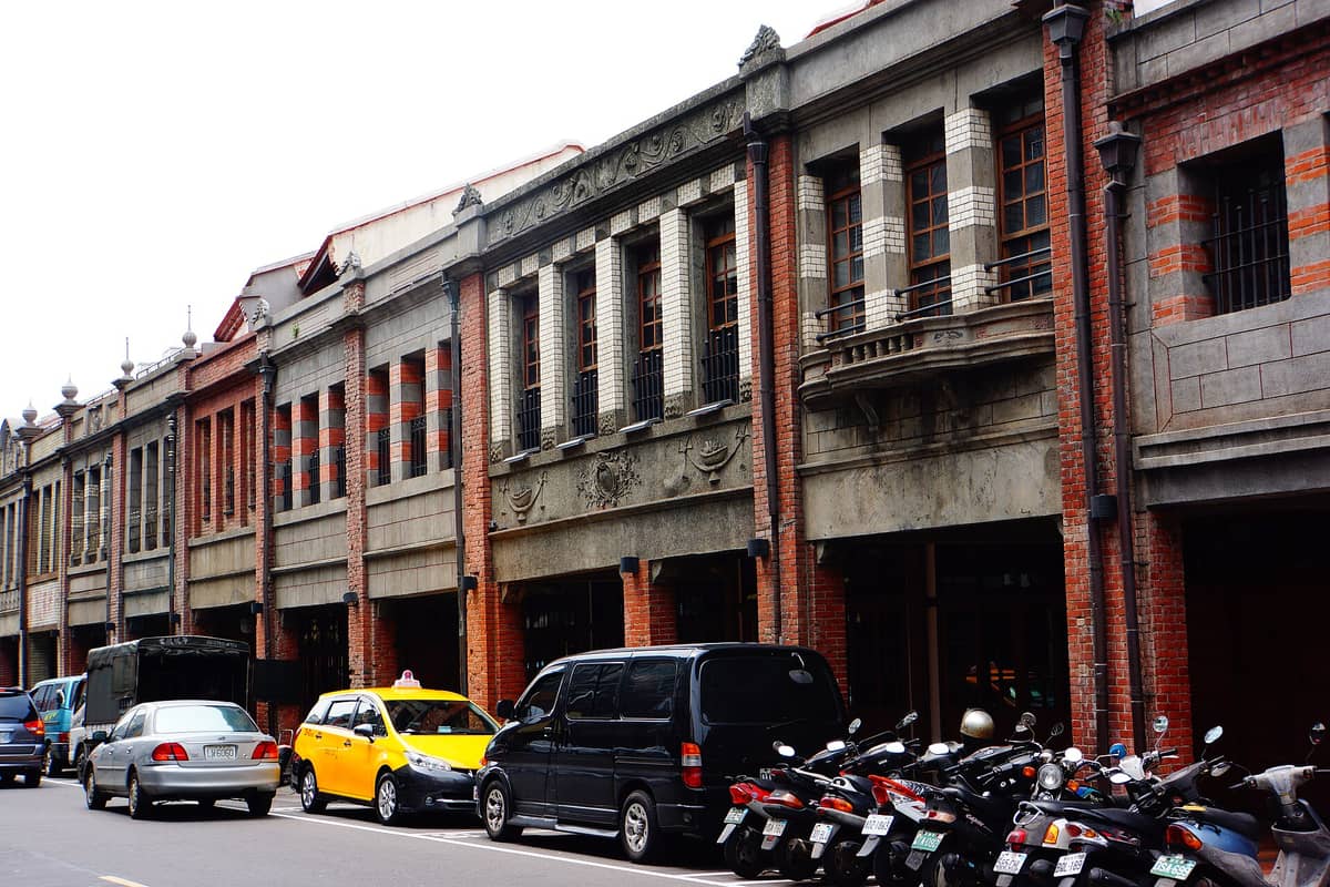Reihen restaurierter Kaufmannshäuser aus roten Backsteinen der Qing-Dynastie entlang des Bopiliao Historischen Viertels in Taipeh, mit geparkten Rollern und Autos am Straßenrand.