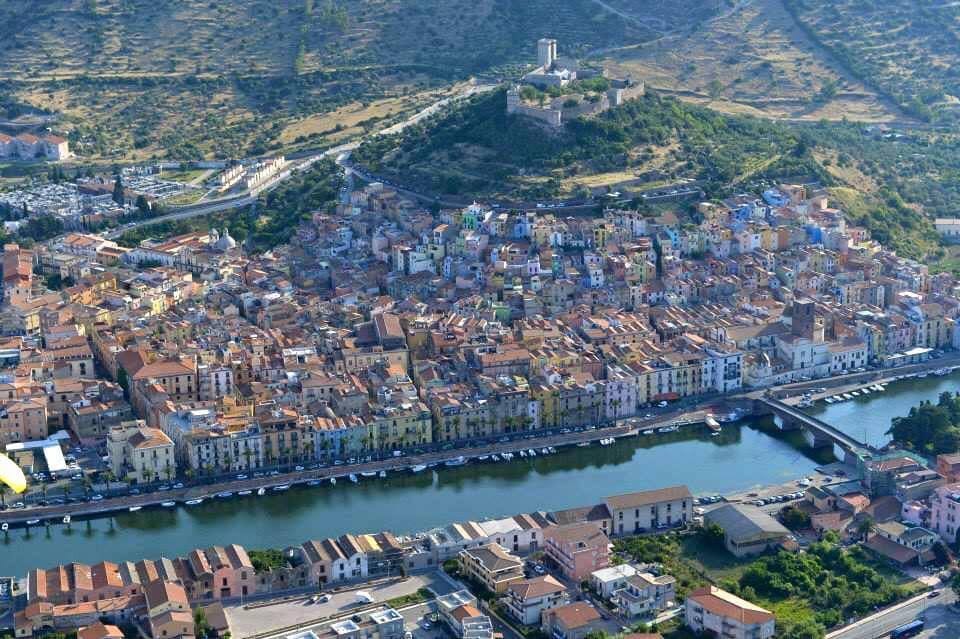 Luftaufnahme von Bosa, Sardinien, mit bunten Häusern am Flussufer, einer mittelalterlichen Burg auf dem Hügel und dem Temo, der durch die Stadt fließt.